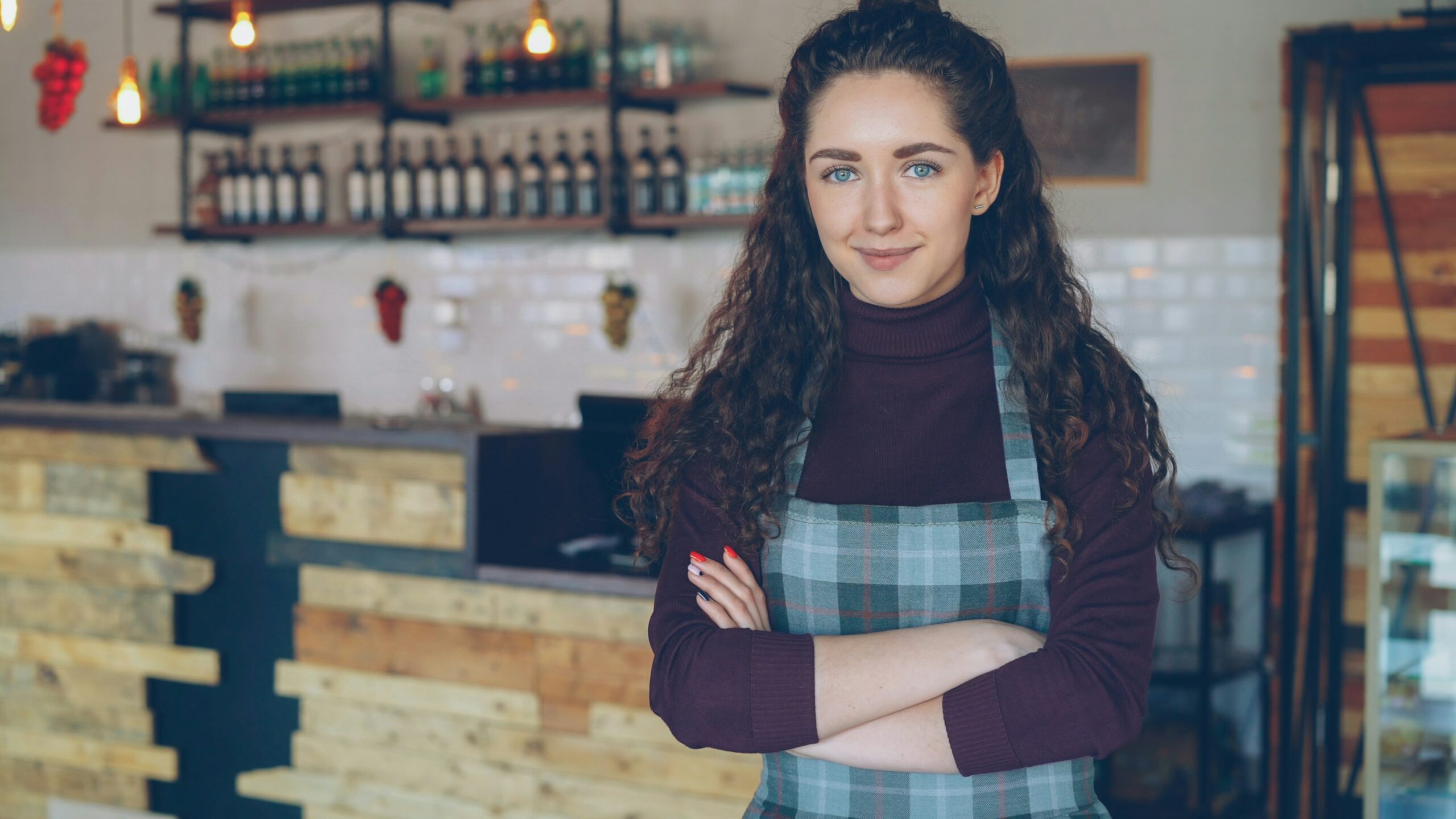 Cafe owenr stands confidently in a checkered apron in front of the cafe counter with her arms crossed.